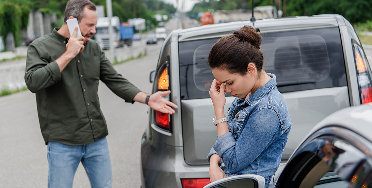 A woman with her hand on her face and a man upset and on his phone in the background after getting into a car accident